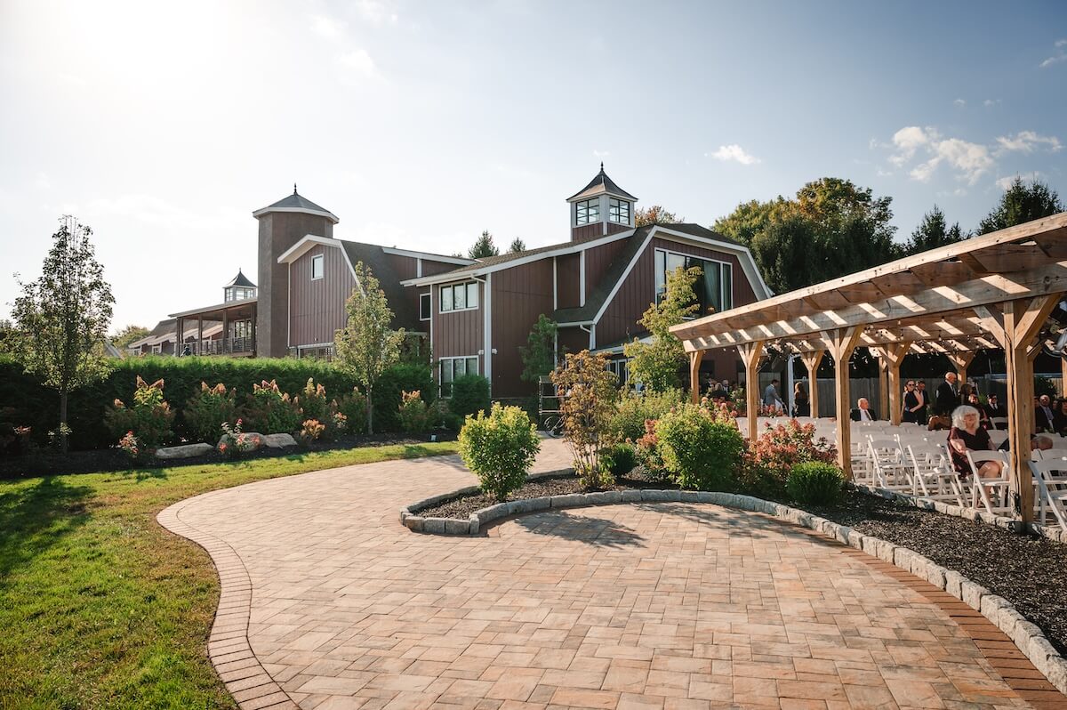 Outdoor photo of rustic Hudson Valley barn wedding venue The Barn at Villa Venezia with pergola outdoor ceremony space on sunny day. Patio leads towards red barn.